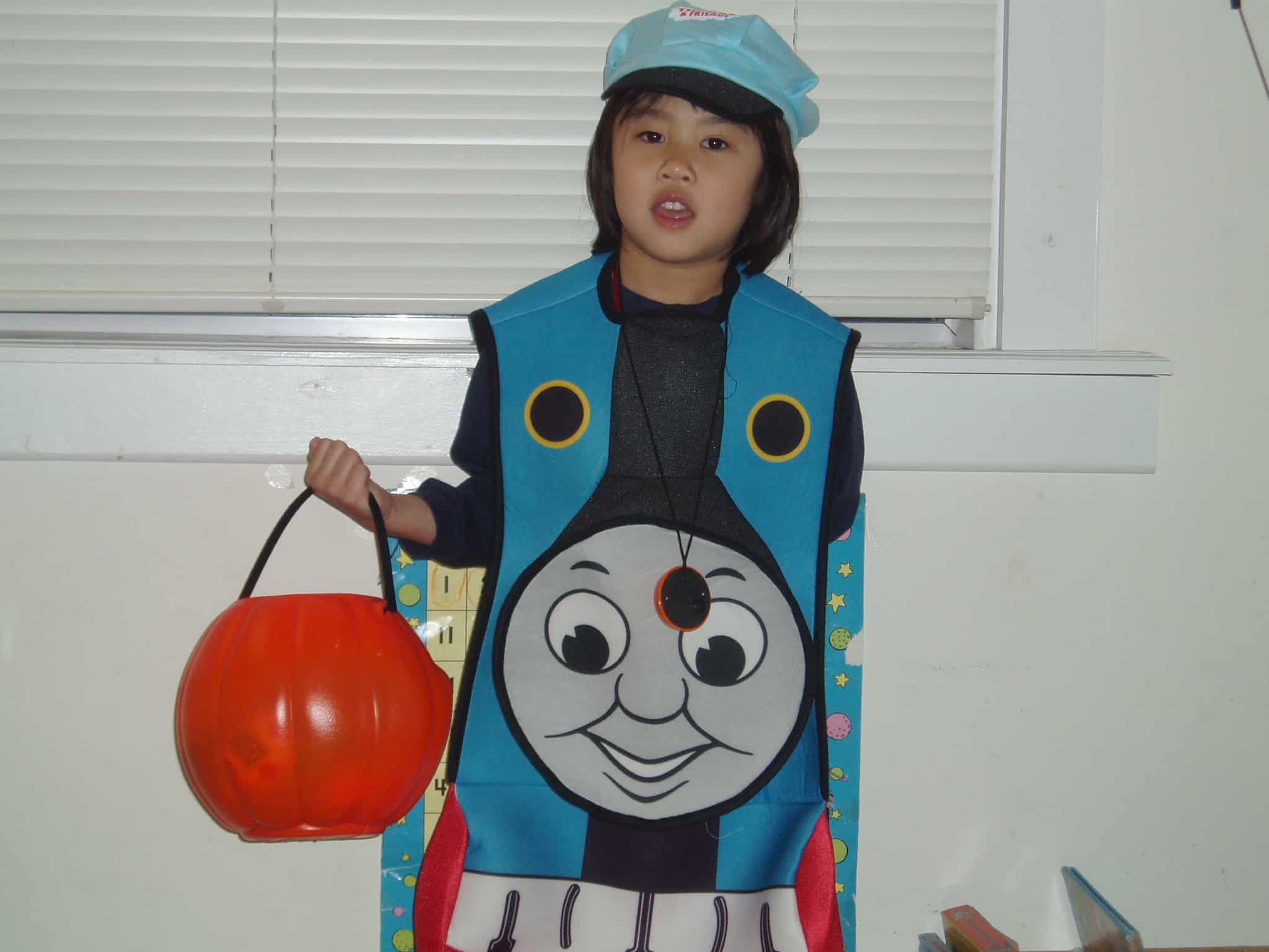 A young toddler Meggie is wearing a Thomas the Train Halloween costume. They are holding an orange pumpkin candy basket. Jefferson City, Missouri, United States of America