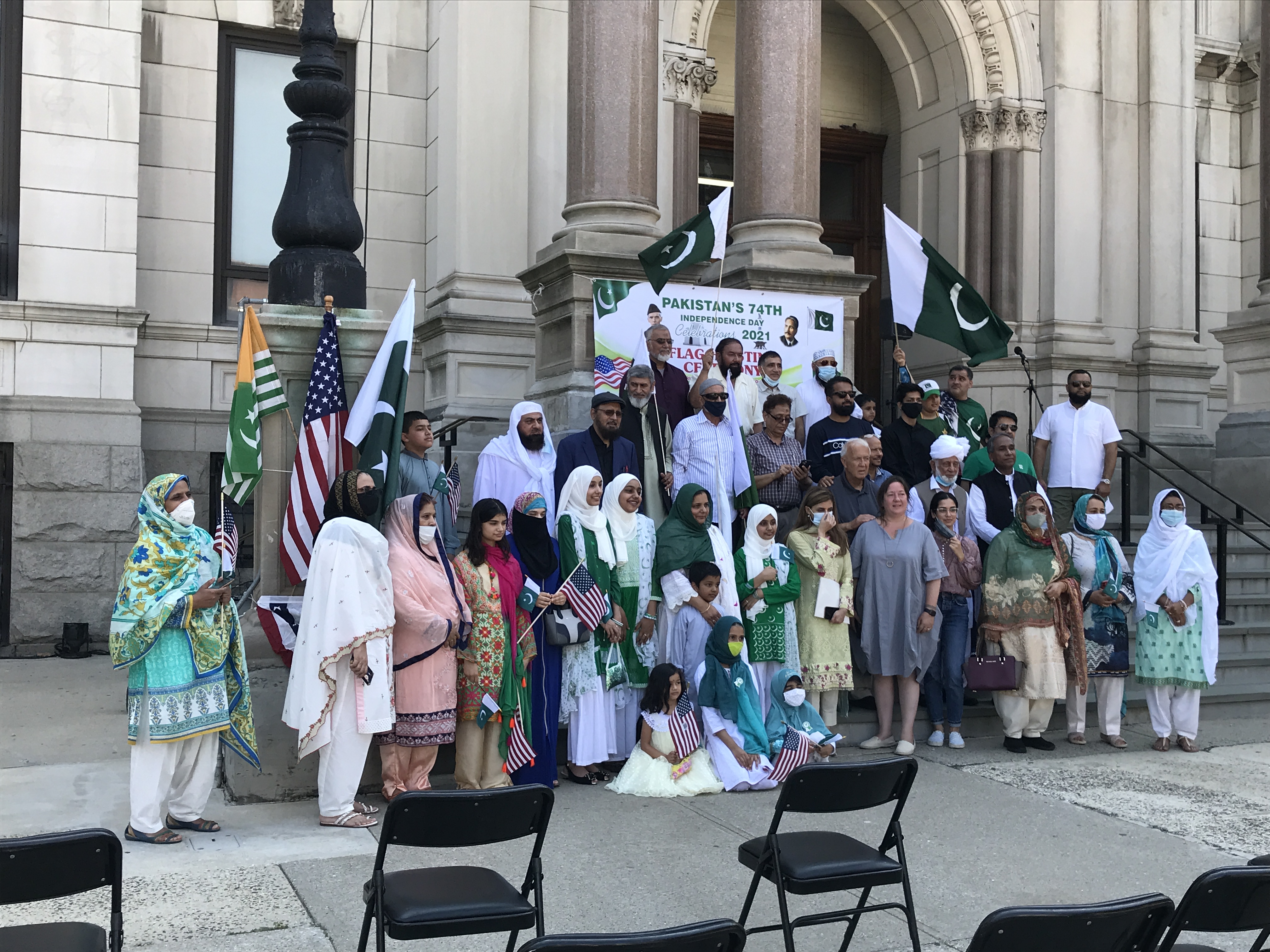 The flag raising ceremony of Pakistan. Many Pakistani Americans are standing together for a group photo. Some of them are wearing green and white clothes, and are holding green and white Pakistani flags. Jersey City City Hall, Jersey City, New Jersey, United States of America