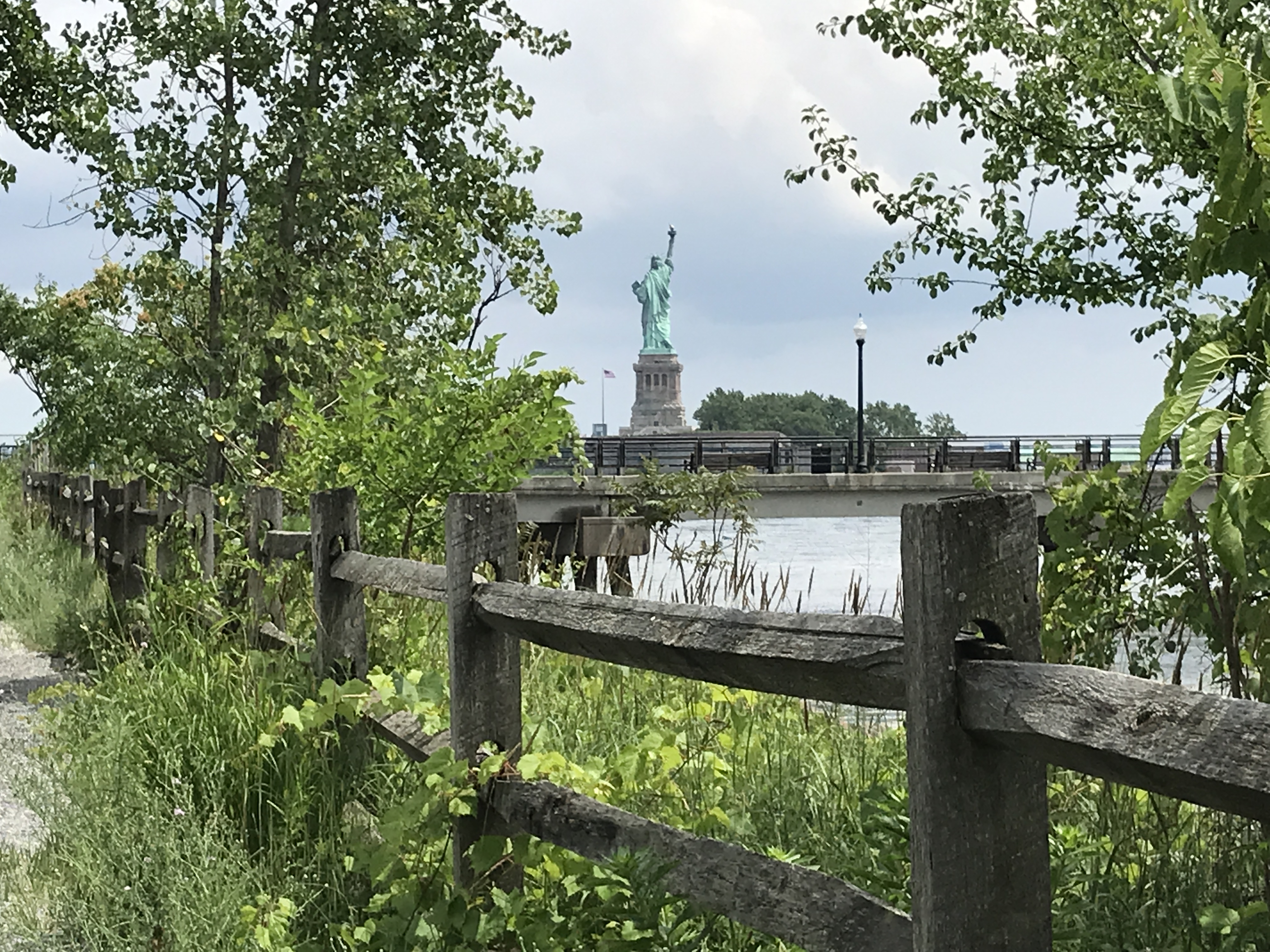 The backside of the Statue of Liberty is juxtaposed with the pond, trees, and fence along a hiking nature trail in Liberty State Park, Jersey City, New Jersey, United States of America