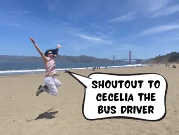 Meggie is wearing a pink blouse and white capri pants, jumping up in the air over a yellow sandy beach with the Golden Gate Bridge in the background. Meggie says in a comic text speech bubble, "Shoutout to Cecelia the bus driver." Baker Beach, San Francisco, California, United States of America.