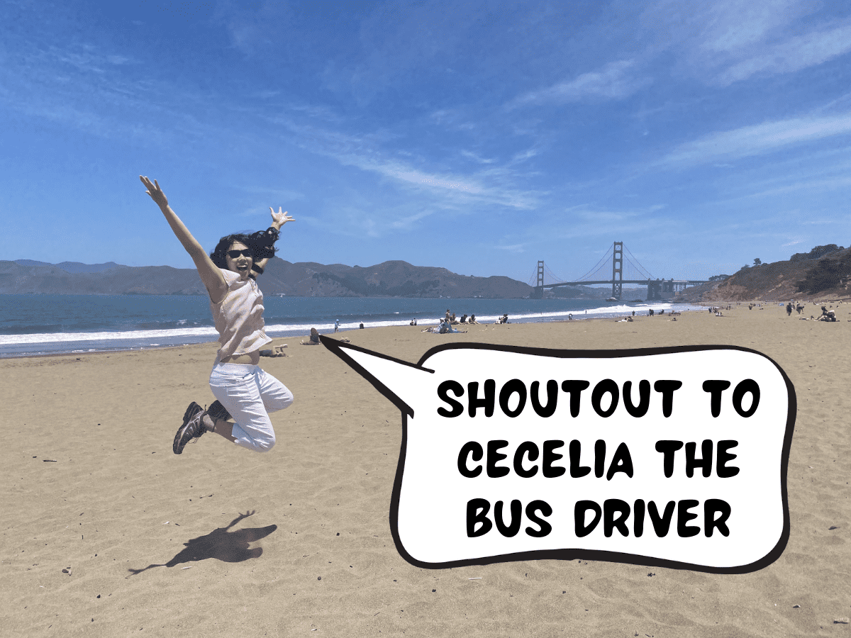 Meggie is wearing a pink blouse and white capri pants, jumping up in the air over a yellow sandy beach with the Golden Gate Bridge in the background. Meggie says in a comic text speech bubble, "Shoutout to Cecelia the bus driver." Baker Beach, San Francisco, California, United States of America.