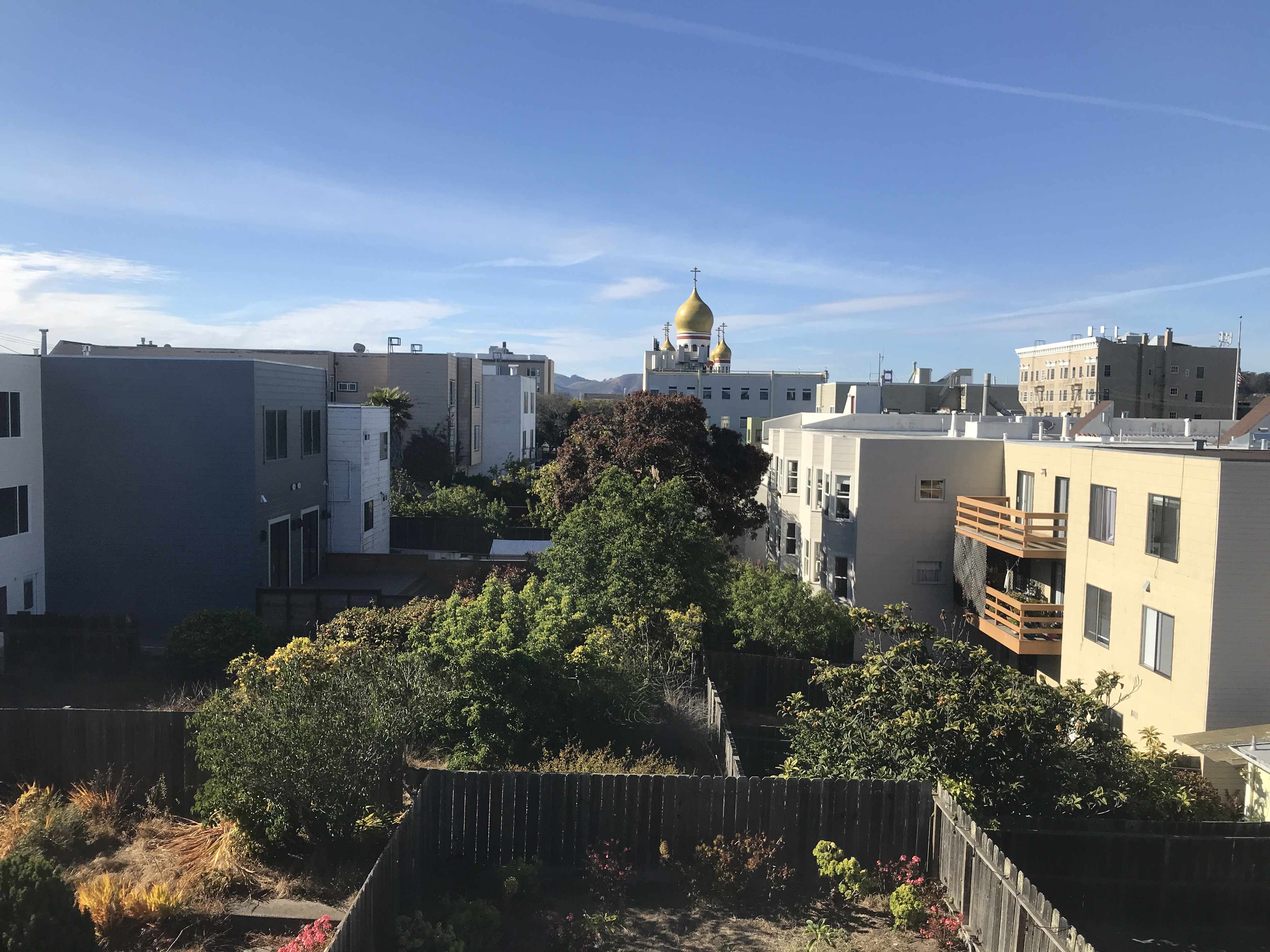 A row of apartments with lots of trees in backyards. A Russian Orthodox church with yellow domes sits in the background. Clear sky with a few small clouds. The Richmond District, San Francisco, California, United States of America.