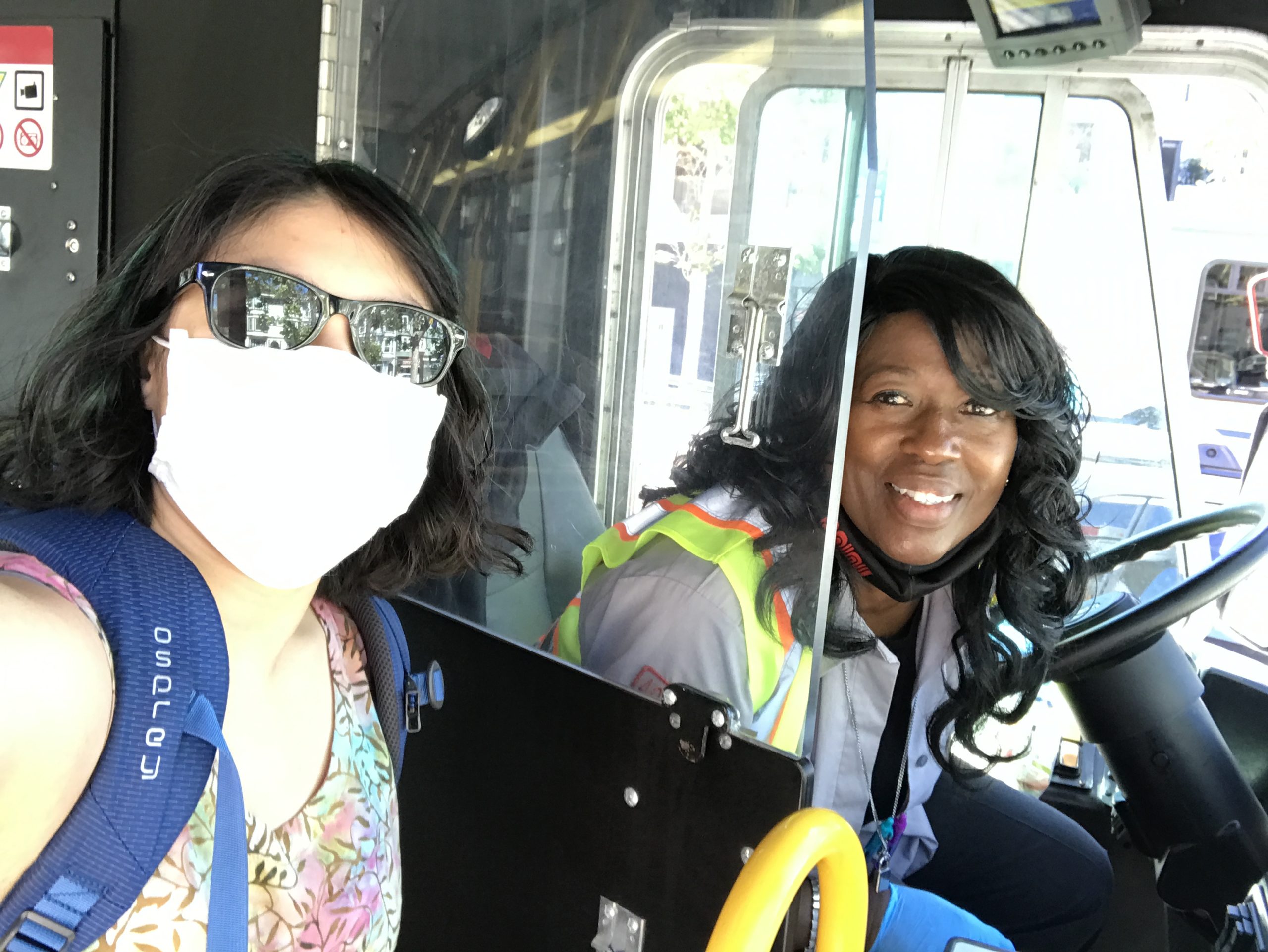 Meggie has a mask on as they pose with Cecilia the bus driver who has a grin on their face and is peeking out through the driver's door. A view from Twin Peaks features a clear blue sky, the blue water of the bay, the white buildings all over the city, and the road leading up to Nearby the Castro District, San Francisco, California, United States of America.