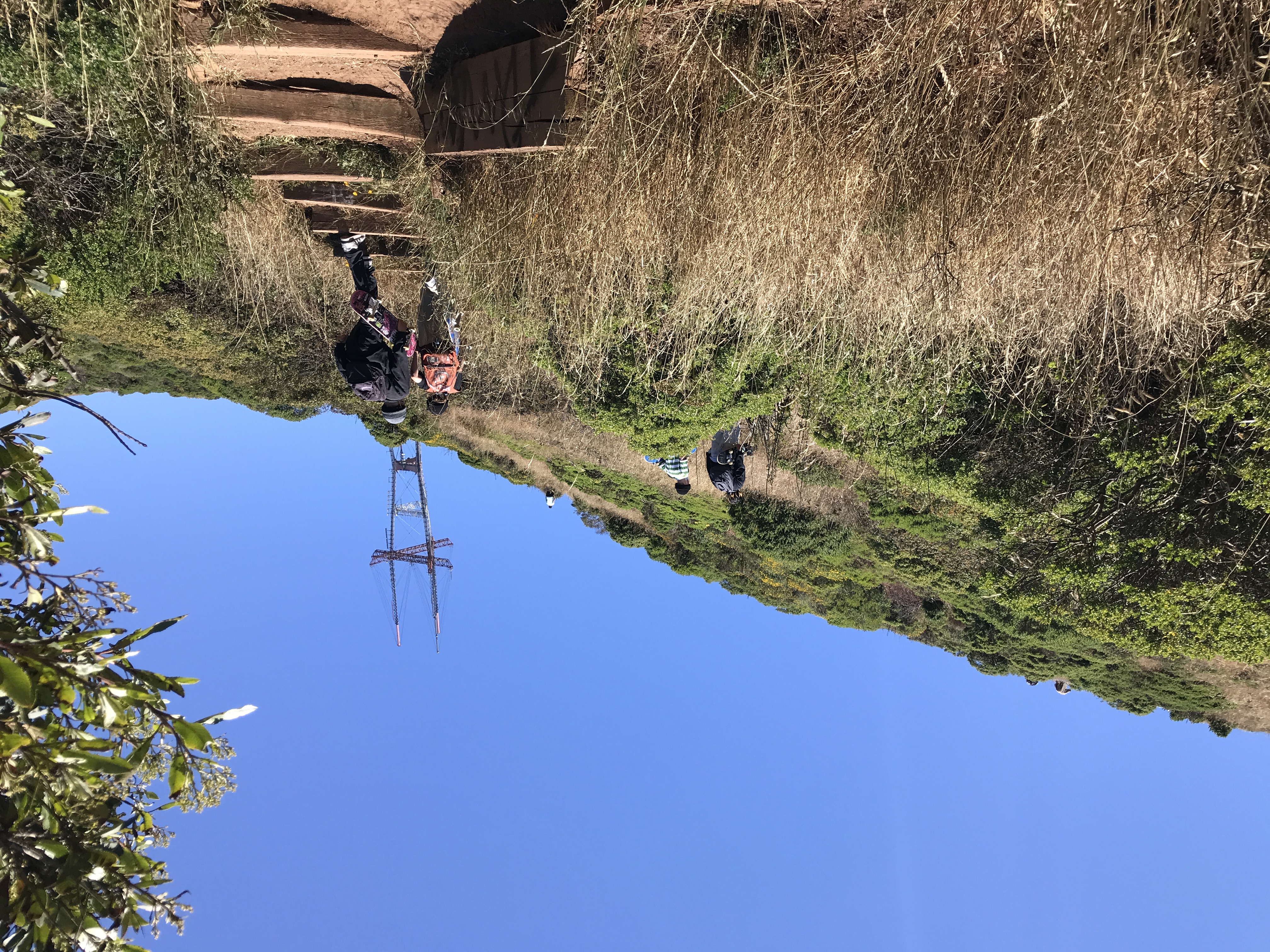 A dirt hiking trail held together with wooden plants goes up a hill alongside yellow and green shrubby bushes and plants. A few skater boys are hiking up the trail under a clear blue sky. Twin Peaks, San Francisco, California, United States of America.