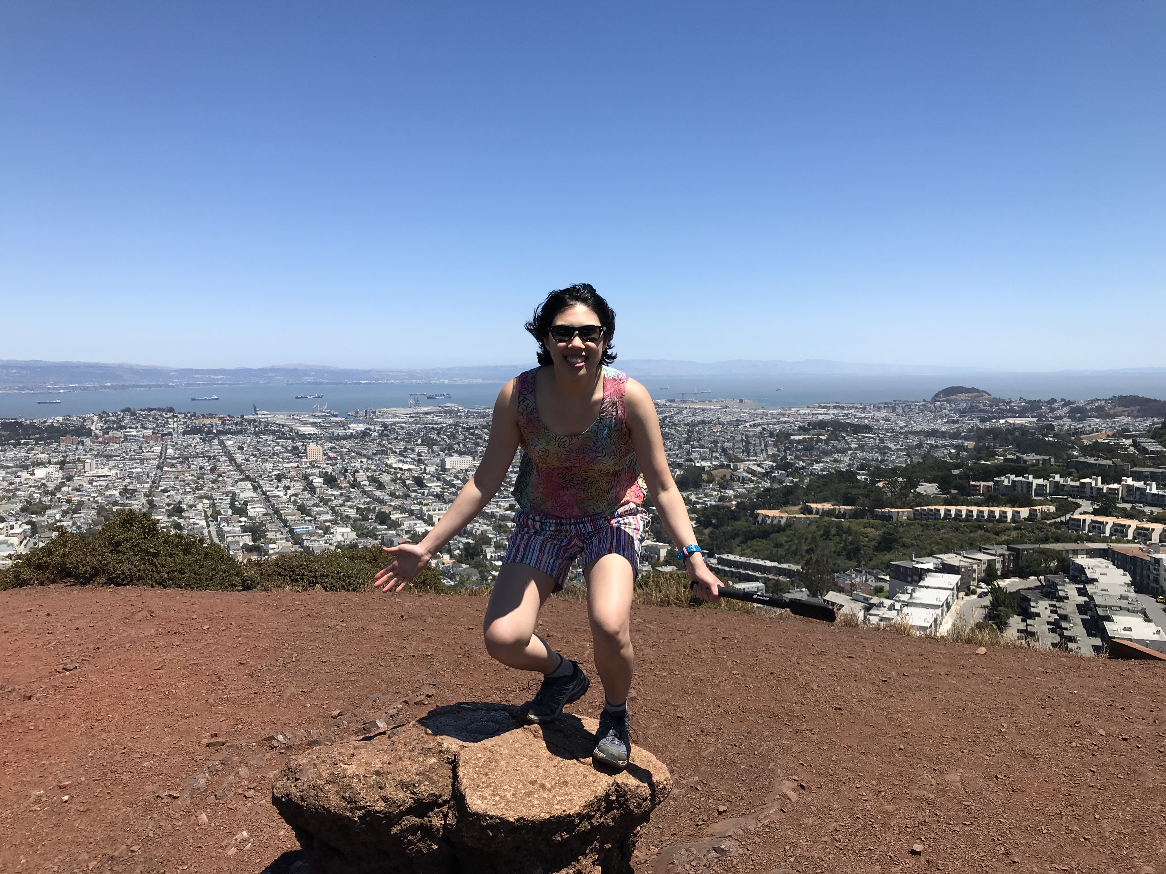 A view from Twin Peaks features a clear blue sky, the blue water of the bay, the white buildings all over the city. Meggie is smiling and standing in front of the view with a colorful shirt with vine patterns. Twin Peaks, San Francisco, California, United States of America.