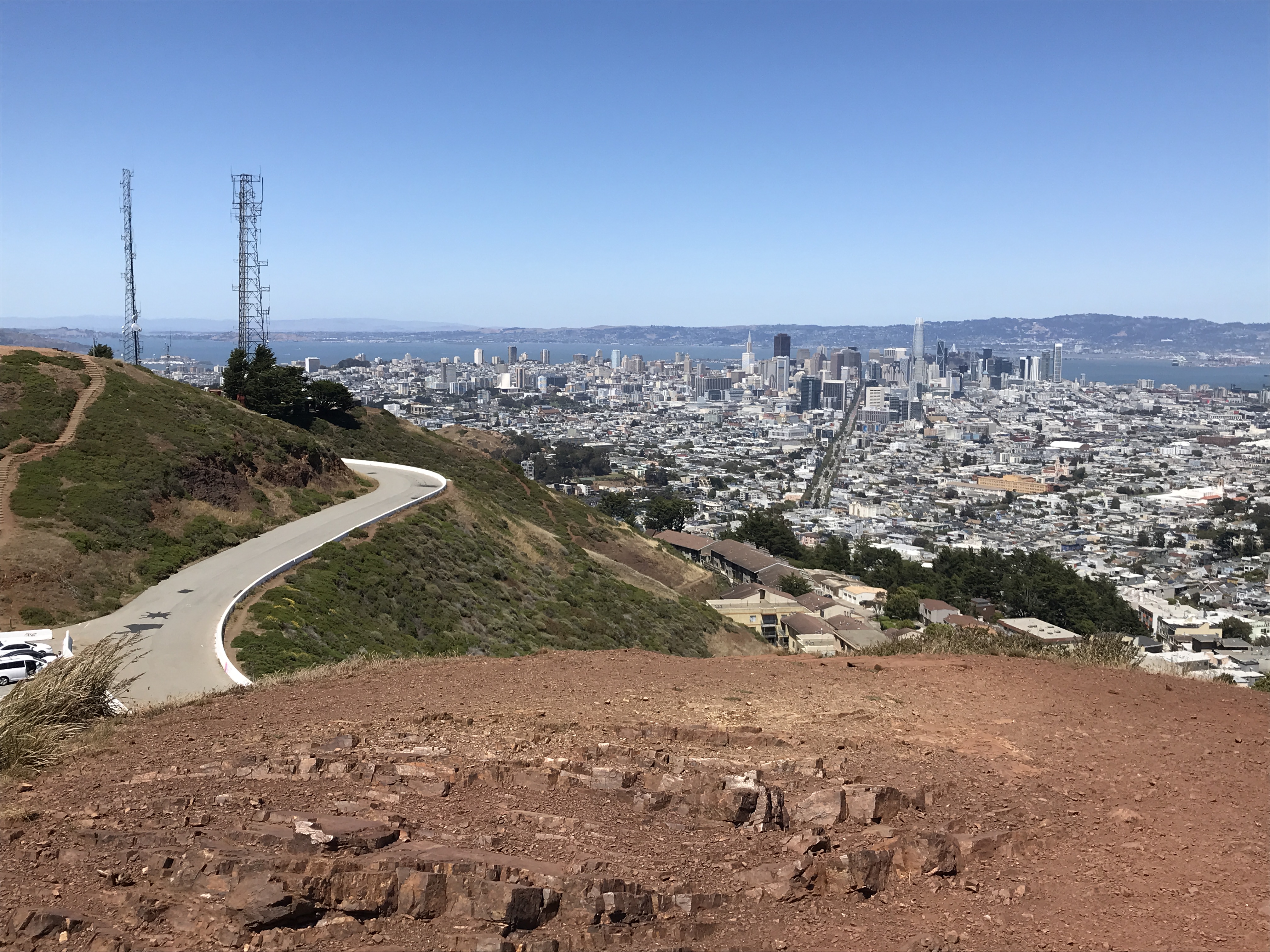 A view from Twin Peaks features a clear blue sky, the blue water of the bay, the white buildings all over the city, and the road leading up to Twin Peaks. Twin Peaks, San Francisco, California, United States of America.