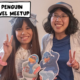 Two penguins from the Club Penguin community, Meggie (Tech70) and Athena (Cw700) meet in real life. Both of them are holding up paper cutouts of their penguins and doing the peace sign with their other hand. Athena is wearing a cute white dress and Meggie is wearing a blue t-shirt and ballcap. Their human outfits have a similar color scheme with their penguin cutout outfits. Jersey City, New Jersey, United States of America
