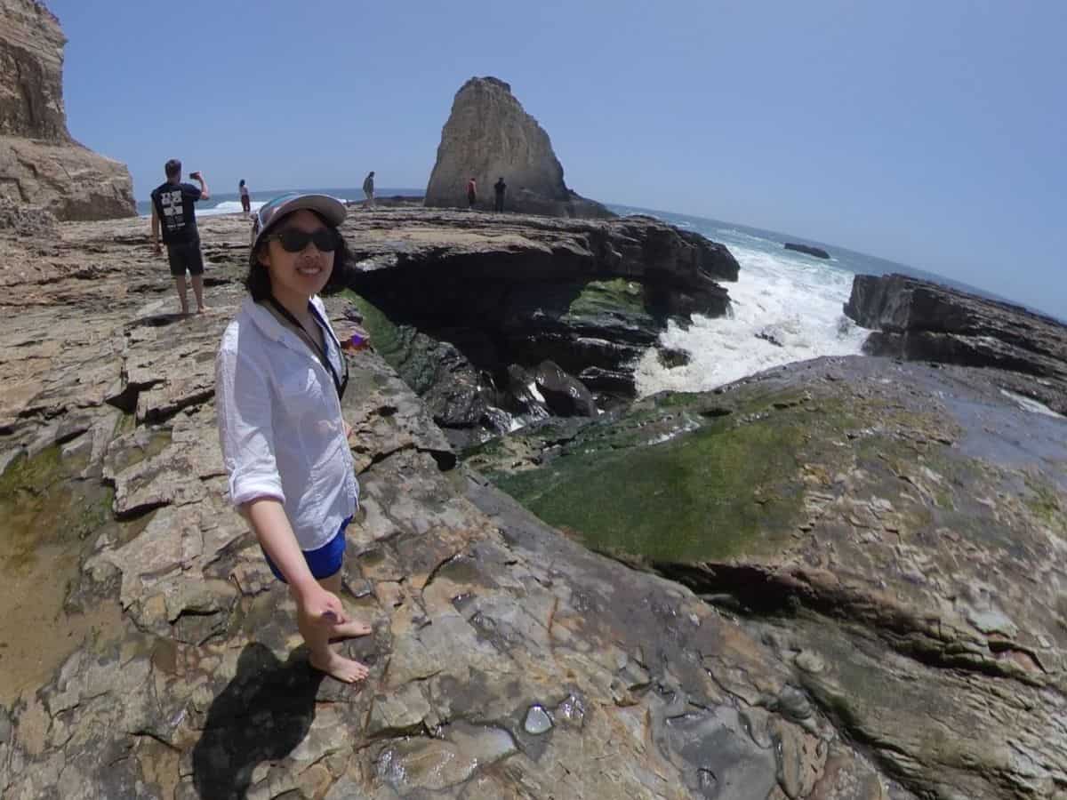 Meggie is smiling on top of the cliff, with the cliffside behind them. Green algae is growing on the rocky surface. The ocean is crashing into the side of the cliff. Sky is blue and clear. Panther Beach, Santa Cruz, California, United States of America.