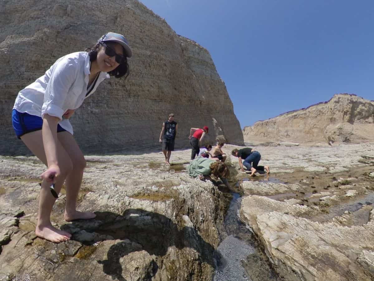 Meggie is smiling as they are standing over a crack in the rocky cliff ground. Many of their friends are crouched down inspecting the crack for sea creatures in the tidepools. Clear blue sky. Panther Beach, Santa Cruz, California, United States of America.