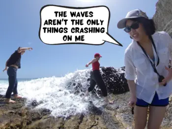 Meggie and her two friends are caught in a surprise wave crashing onto them. A clear blue sky and brown cliffs and rocky grounds. Meggie, in a comic text bubble, is saying, "The waves aren't the only things crashing on me." Panther Beach, Santa Cruz, California, United States of America.