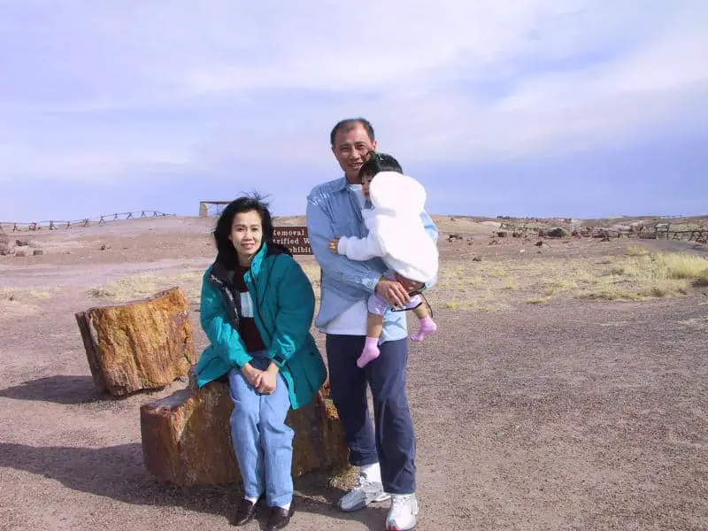 Meggie's dad is holding baby Meggie. Meggie's mom is sitting on a petrified log. In the background is a bare desert with scattered petrified logs. Petrified Forest National Park, Arizona, United States of America.