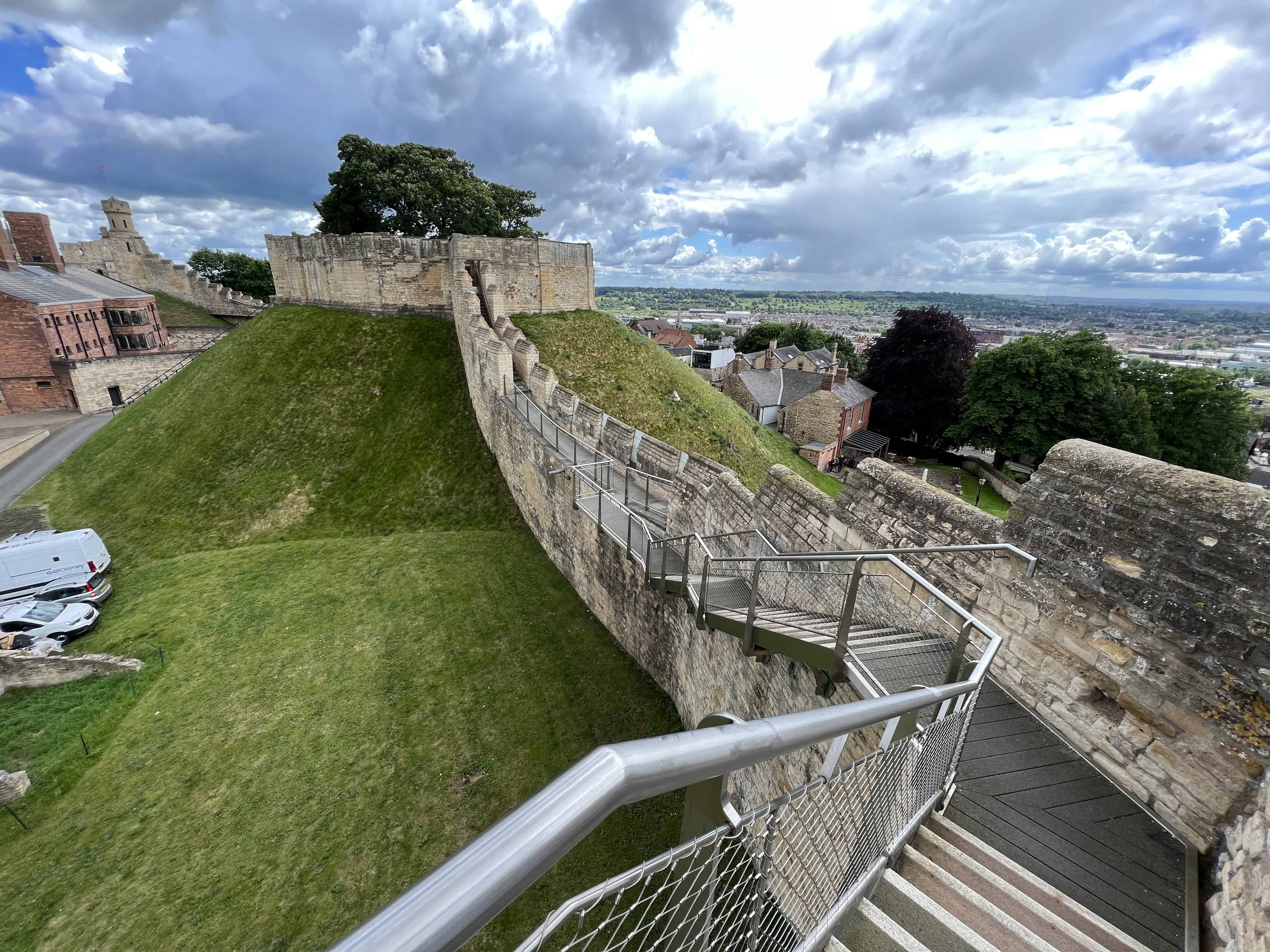 The tan, stained with black tarnish, castle walls running along the green grassy hills. The red-bricked buildings of the town of Lincoln are outside the walls. On the grounds of Lincoln Castle, Lincoln, Lincolnshire, England, United Kingdom.