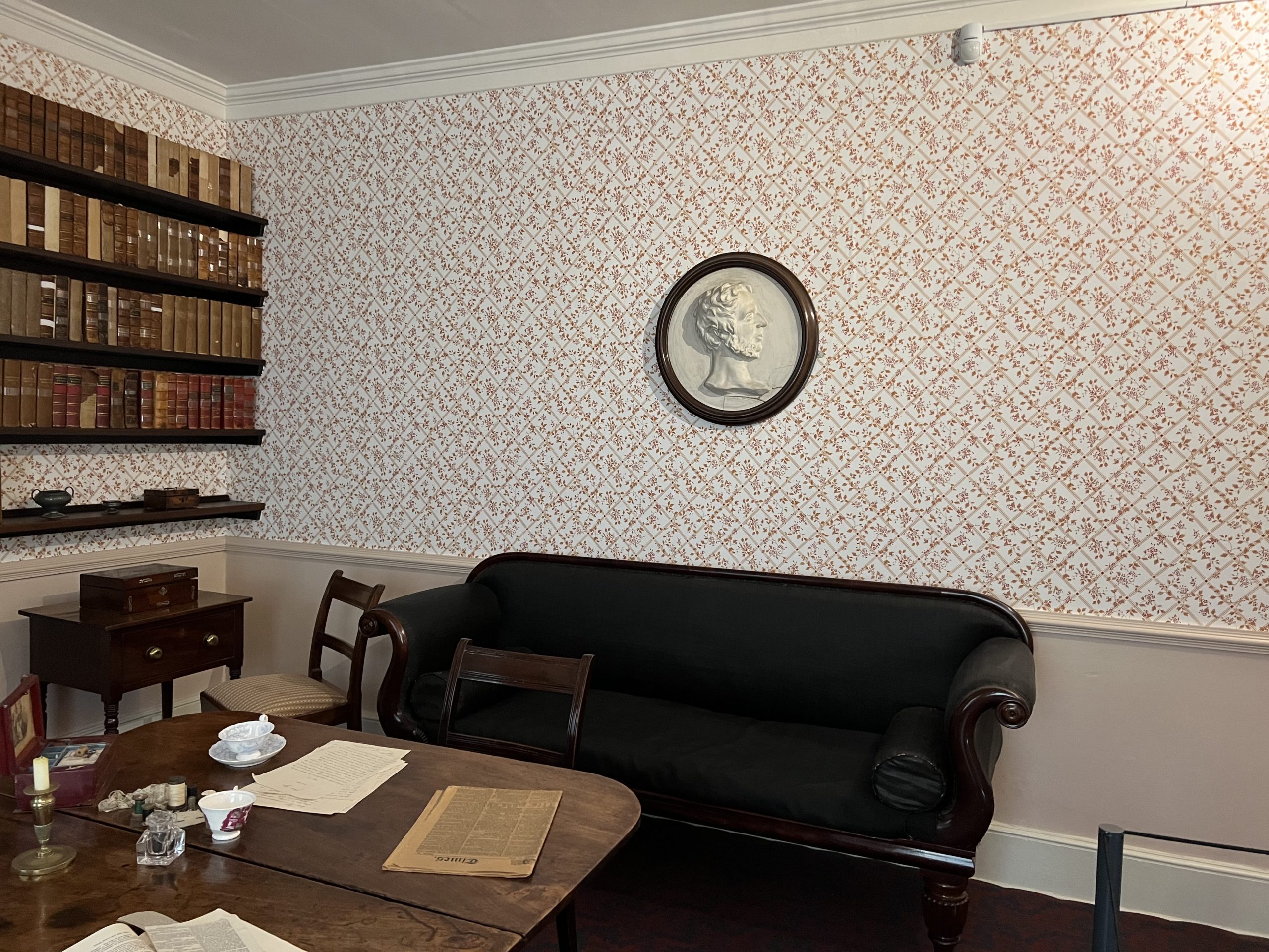 The black sofa in the Brontë Parsonage's dining room. The wallpaper has red flowers. There is a wall shelving of many old brown and red books. Brontë Parsonage, Haworth, Bradford, West Yorkshire, England, United Kingdom.