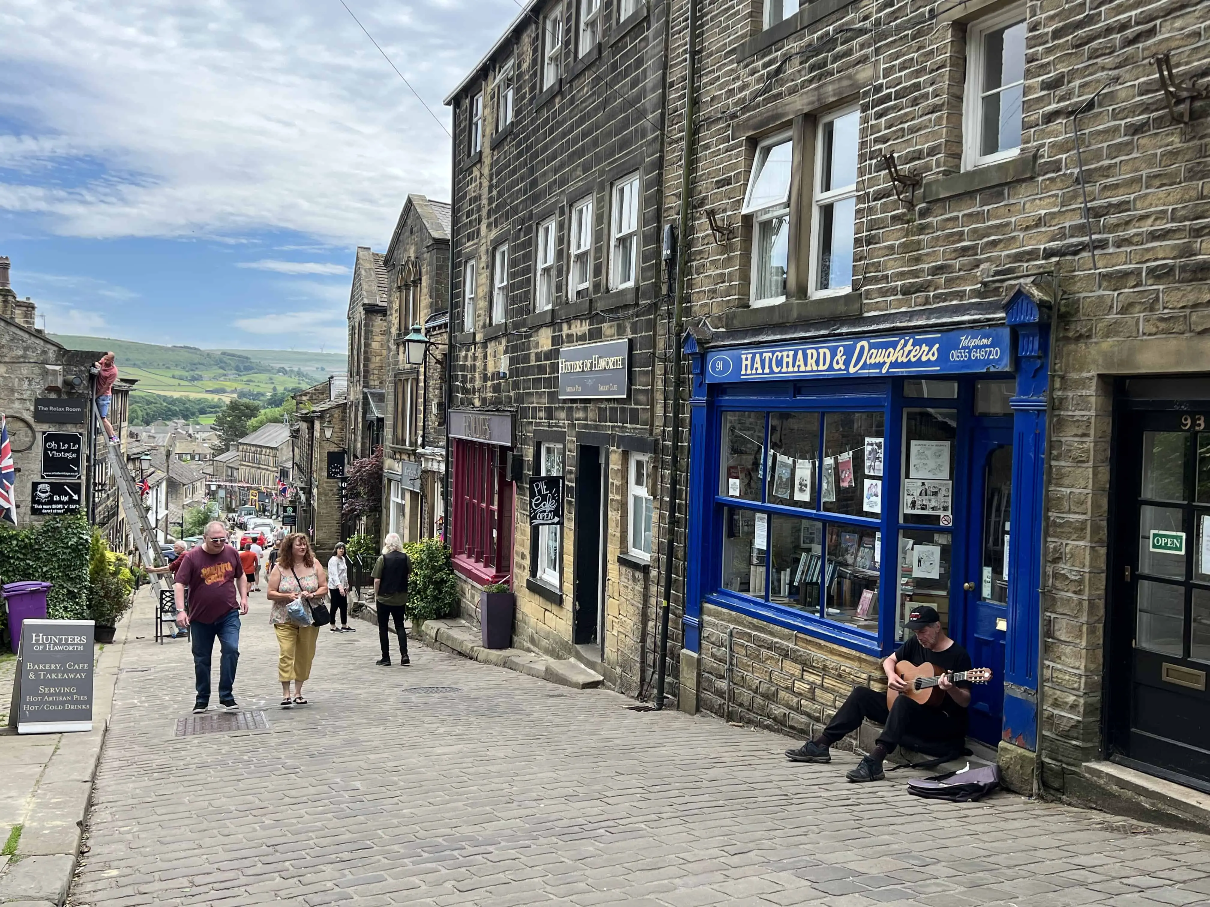A man with a classical guitar is sitting at a storefront to collect donations. Haworth's main street has black and tan bricked-buildings and a gray cobblestone street. The rolling green hills and trees of the English countryside is in the distance. Main Street, Haworth, Bradford, West Yorkshire, England, United Kingdom.