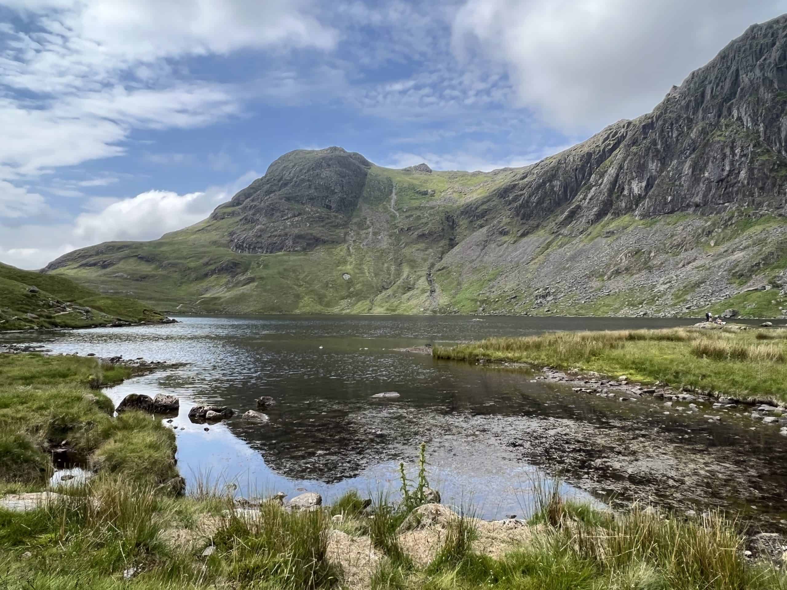 A blue lake surrounded by green grass. A gray mountain range hovers over the lake. Lake District National Park, England, United Kingdom.