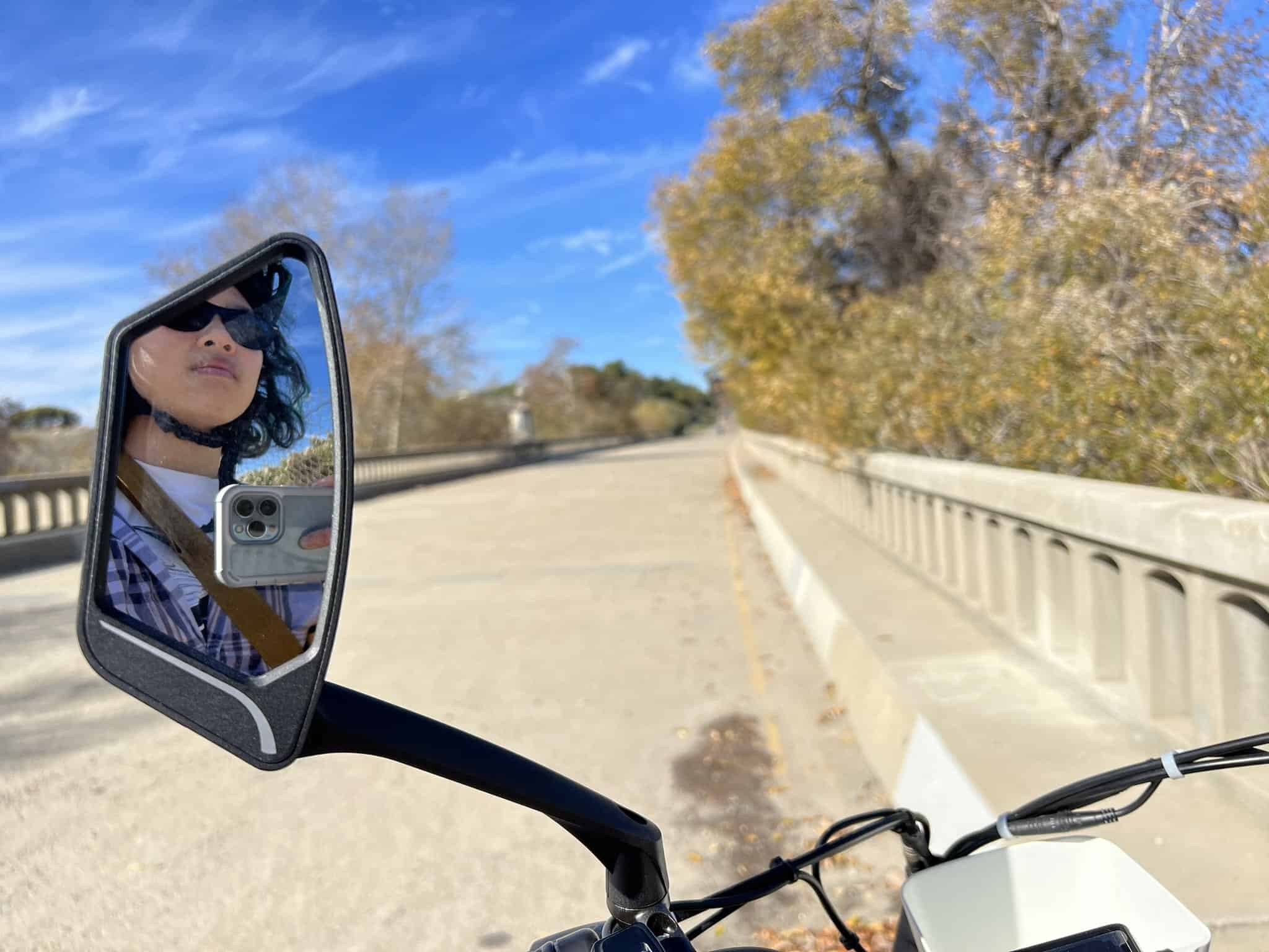 A highway with yellow trees on the side. A bicycle’s handlebars in the front of the picture. A bicycle’s rearview mirror with a reflection of Tech70/Meggie wearing blue plaid.
