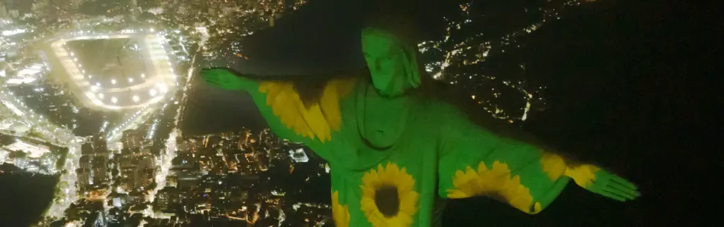 The huge Christ the Redeemer statue in Rio de Janeiro, Brazil, at night. It is light projected with yellow sunflowers against a green background.