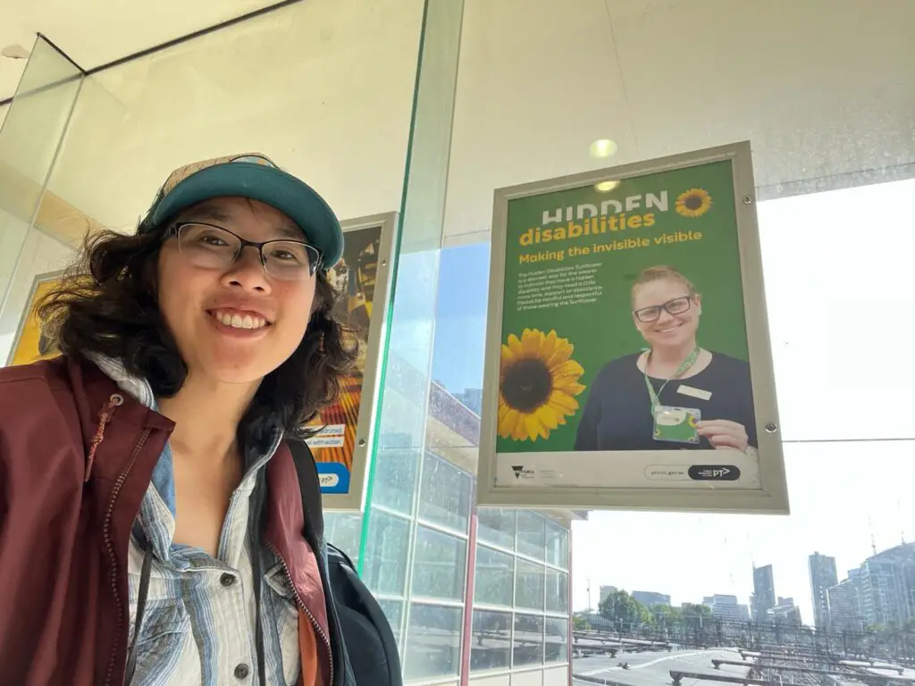 Meggie in a maroon jacket taking a selfie with a green Sunflower Lanyard poster at Flinders Street Railway Station, Melbourne, Australia.