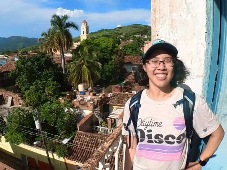 Meggie is wearing a Daytime Disco shirt on the top of a bell tower in Trinidad, Cuba. Green trees and historic buildings with brown roof tops are seen below the tower.