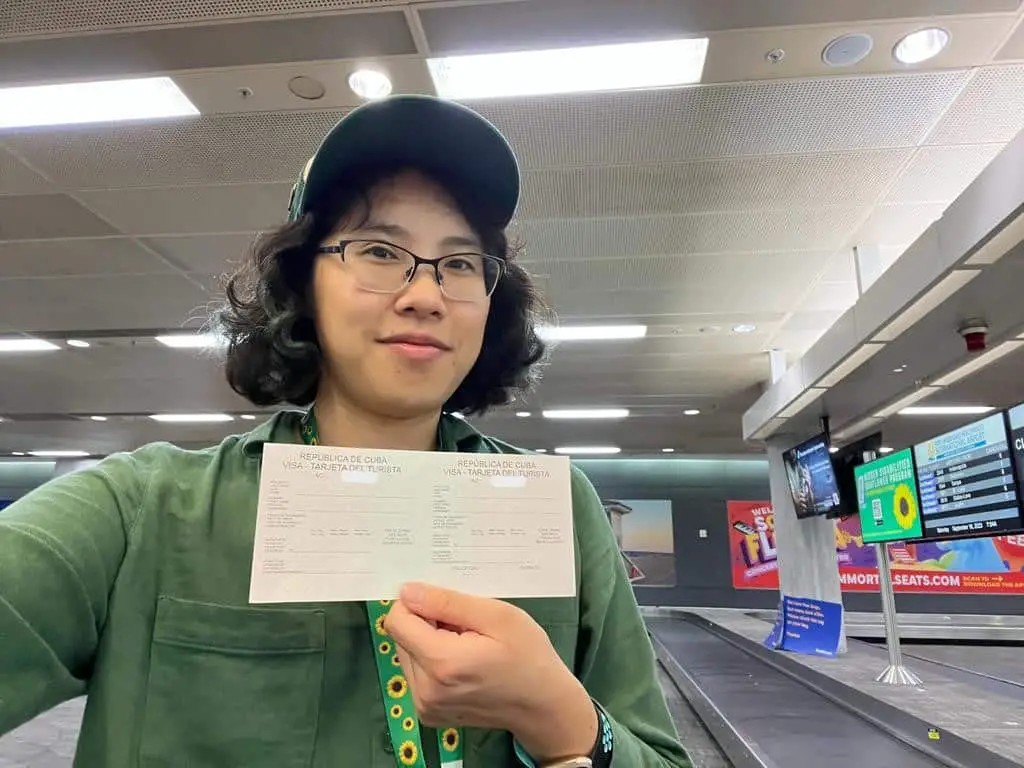 Meggie in a green jacket is holding up the pink Cuba visa at Fort Lauderdale Airport.