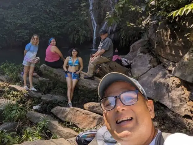 Meggie, their tour mates, and tour leader at a secret waterfall pooling into a big pond surrounded by boulders and green plants.