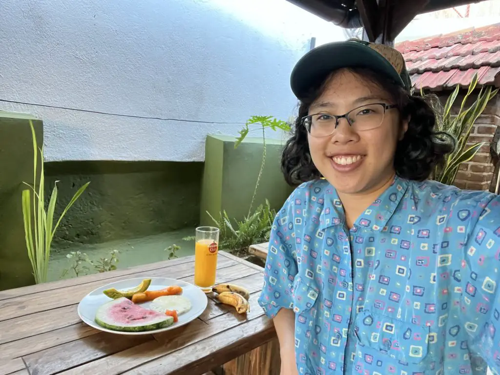 Meggie in a blue shirt eating at a breakfast table with a plate of fruit.