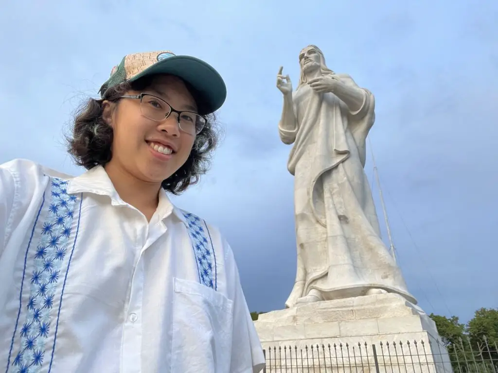 Meggie in a handmade blue Cuban shirt is standing in front of the white statue of the Christ of Havana.