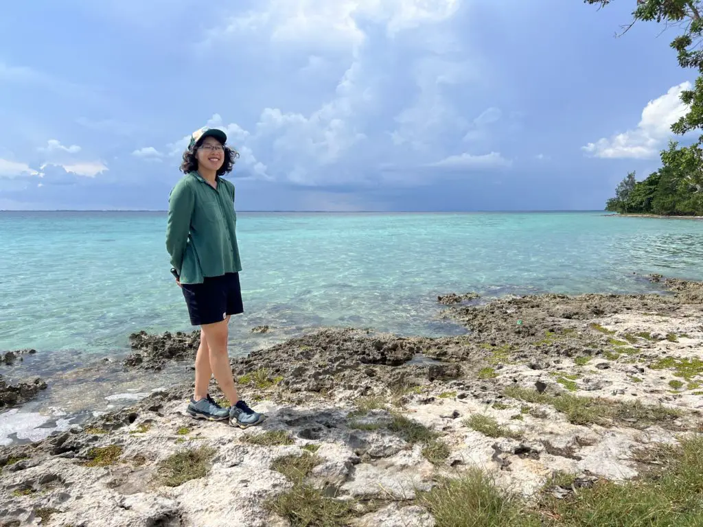 Meggie standing near the clear turquoise waters at the Bay of Pigs in Cuba. Meggie is wearing a green shirt and black shorts.