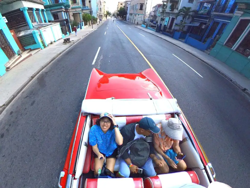 Meggie in a blue shirt is riding in a classic red car through the colorful blue streets of Havana with their tour leader