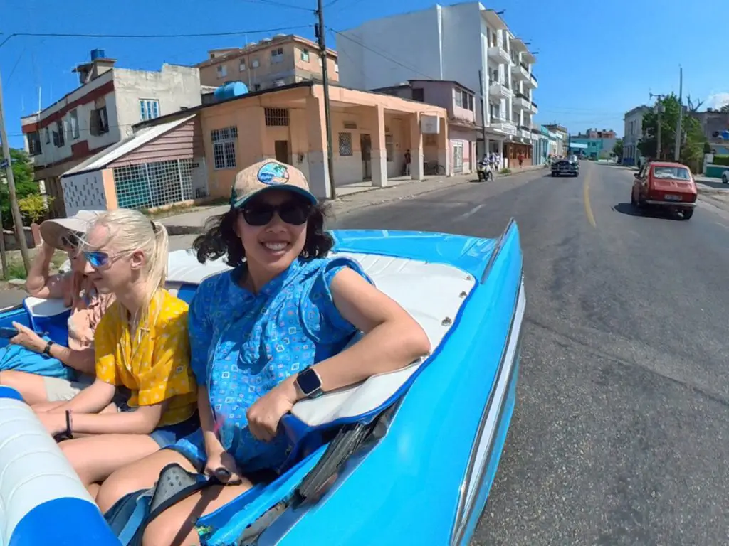 Meggie in a blue shirt riding in a classic blue 1950s American convertible with two other tour group members. The streets of Havana have other older cars and some colorful, older buildings.