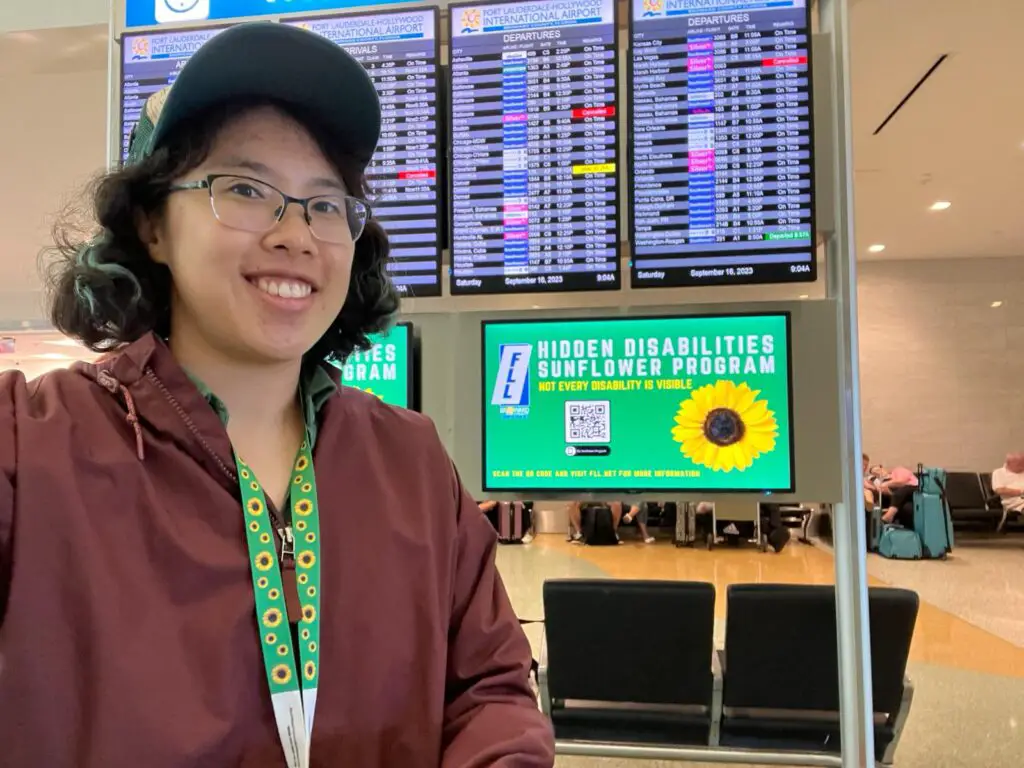 Meggie in a maroon jacket wearing a green Sunflower lanyard and standing in front of a green Sunflower Lanyard TV monitor next to the airplane arrival and departure screens in Fort Lauderdale-Hollywood International Airport.
